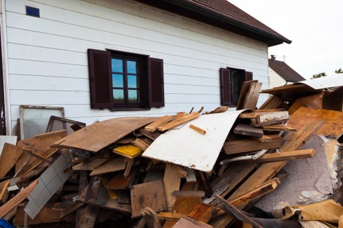 A large pile of mixed construction and household waste materials, including wooden planks, particle boards, and cardboard, is stacked against the side of a white residential building with dark brown window shutters in Woolwich. The debris appears to have been recently gathered and is situated on a paved driveway or yard area, with some pieces leaning or partly resting on each other, creating a disorganized heap. The wooden items show various textures and finishes, from smooth painted surfaces to rough cut edges, with some pieces displaying nails or fasteners. The background reveals part of the house's exterior wall and roof, under natural daylight, suggesting an outdoor rubbish removal scene typical of property clearances in the Woolwich postcode district. This image illustrates waste collection efforts undertaken by companies like Office Clearance Woolwich, specialists in domestic and commercial rubbish clearance services.