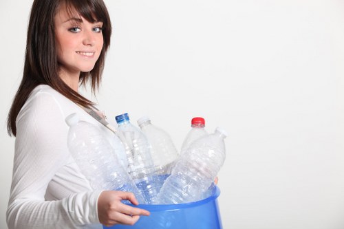 A young woman with shoulder-length brown hair, wearing a white long-sleeved top, is holding a blue plastic bin filled with empty, clear plastic bottles with different coloured caps. The bottles are transparent, some with visible ridges, and appear lightweight. She stands against a plain white background, smiling slightly and looking at the camera. The scene is well-lit, emphasizing the crisp clarity of the bottles and the smooth surface of the plastic bin. This image suggests a focus on recycling or waste collection, relevant to rubbish removal services such as those offered by Office Clearance Woolwich, who operate in Woolwich postcode areas. The composition highlights the process of gathering recyclable plastics, suitable for website content related to rubbish clearance or waste management in the Woolwich locality.