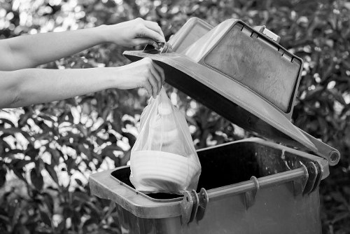 A person is disposing of a clear plastic bag filled with white ceramic items into a black rubbish bin with a lid attached to a curbside on a driveway. The bin is made of durable plastic, featuring a textured surface and sturdy handles, positioned against a backdrop of leafy shrubbery. The lid is partially open, revealing the bag being lowered, with the person's arms visible, one holding the bag and the other opening the lid. The scene suggests an outdoor waste disposal setting typical for local rubbish removal services like those offered by Office Clearance Woolwich, serving residential areas in Woolwich or nearby postcode zones. The natural light enhances the contrast between the dark bin and the lighter bag, emphasizing the activity of waste disposal in a tidy, environmentally conscious manner within a suburban environment. The image captures a routine part of rubbish collection, highlighting the importance of proper waste management and recycling at the curbside.
