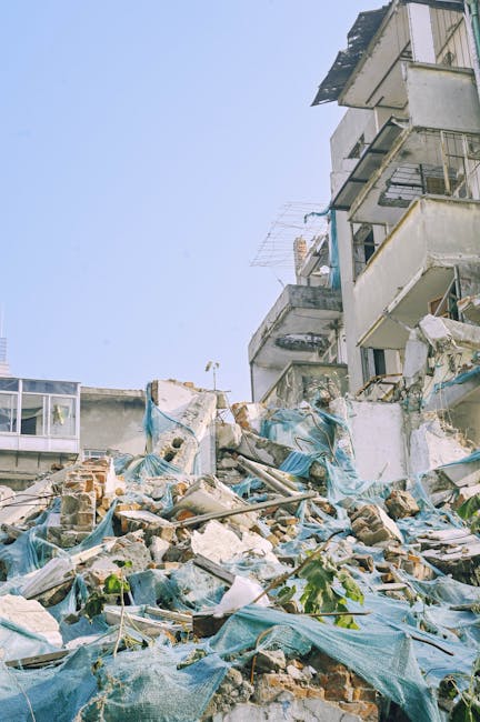 A pile of mixed construction debris and building rubble situated on a paved surface adjacent to a partially demolished brick wall and a white plastered wall, with scattered red bricks, broken white in