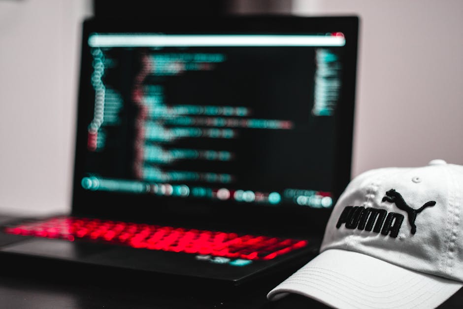 A close-up of a black laptop with a red-backlit keyboard displaying lines of computer code on its screen, positioned on a dark surface. To the right of the laptop, a white Puma-branded baseball cap wi