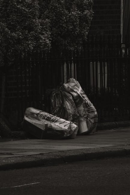 Three large black plastic bags filled with rubbish are situated on a pavement near a black wooden fence, with one bag leaning against a tree and the other two positioned close together. The bags appea
