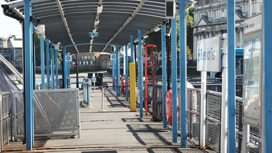 A ferry terminal platform with a long, covered walkway featuring blue metal support posts and a corrugated metal roof. The concrete surface is partially shaded with shadows cast by the overhead struct
