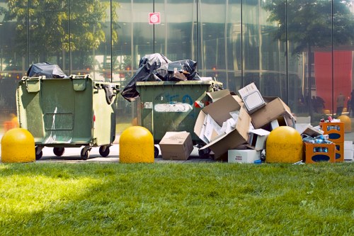 A row of three large waste bins on a paved area adjacent to a grass verge, with the first two bins being green with black lids, and the third bin missing its lid and overflowing with cardboard boxes, plastic bottles, and miscellaneous rubbish. The waste materials include flattened cardboard boxes, plastic containers, and bags, some of which are draped over the bins and scattered on the ground. Behind the bins, there is a reflective glass building face with trees and street furniture partially visible through the glass, under bright daylight. Bright yellow concrete bollards border the grassy area, emphasizing the location as part of a commercial or public space, consistent with rubbish collection and disposal in Woolwich. The scene is typical of a waste management scenario where rubbish from offices or shops is temporarily stored before collection by a waste removal service, such as Office Clearance Woolwich, ensuring the clearing of accumulated waste in urban environments.