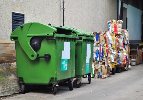 A bright green, large wheeled rubbish bin with a closed lid, positioned on an urban pavement next to a beige building wall. To the right of the bin, there are several bulky stacks of flattened cardboard boxes, mostly brown with some coloured packaging and printed labels, which are leaning against the wall. The ground is paved with grey concrete, and there is a small section of a brick wall visible in the background as well as a black vent or louvre near the building's surface. The scene appears to be part of a commercial or residential area where waste collection and disposal are managed locally, reflecting typical rubbish removal services provided by Office Clearance Woolwich in the Woolwich postcode area. The image captures a cleanup or waste collection site, with the cardboard awaiting disposal or collection, and the overall environment is characterized by a utilitarian and urban atmosphere suitable for professional rubbish removal operations.
