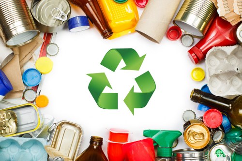 A variety of discarded plastic bottles, aluminum cans, cardboard tubes, and other mixed waste items are scattered on a white surface, with some items partially overlapping. Prominently displayed in the center is a green recycling symbol with three arrows forming a triangle, indicating recycling efforts. The waste includes bottles of different colors such as brown, red, blue, and yellow, alongside crushed cans and flattened cardboard. The background appears clean and well-lit, suggesting a focus on waste separation and recycling practices. This scene represents the type of rubbish materials that companies like Office Clearance Woolwich handle when providing rubbish removal services to commercial or local clients in Woolwich and surrounding areas, highlighting the importance of waste segregation and environmentally conscious disposal.