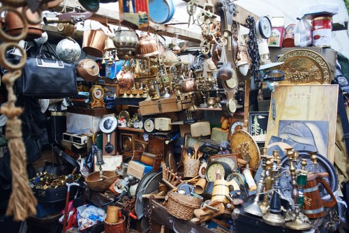 A densely packed outdoor display of various household and decorative items, including metallic ornaments, brass and copper containers, picture frames, clocks, and small furniture pieces, all arranged on multiple shelves and tables. The items vary in shape, size, and finish, with some exhibiting shiny polished surfaces while others appear aged or tarnished. In the background, more objects fill the space, suggesting a cluttered environment typical of a second-hand market or antique stall, possibly located in an area similar to Woolwich. The scene includes a mix of metals, ceramics, and wooden items, some stacked or leaning against each other, forming a visually busy and eclectic arrangement. This images aligns with the type of rubbish that Office Clearance Woolwich might encounter when clearing such clutter from commercial or residential premises, highlighting the need for professional waste removal services in the Woolwich area.